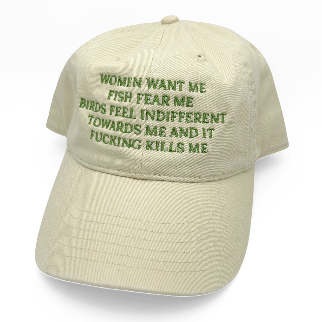 A light beige baseball cap with green embroidered text on the front that reads, 'WOMEN WANT ME FISH FEAR ME BIRDS FEEL INDIFFERENT TOWARDS ME AND IT FUCKING KILLS ME.'.